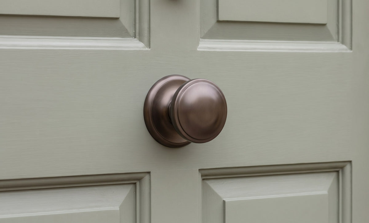 Matt bronze stepped centre door knob on a sage green panelled front door, oak frame and honey Cotswold stone beside it