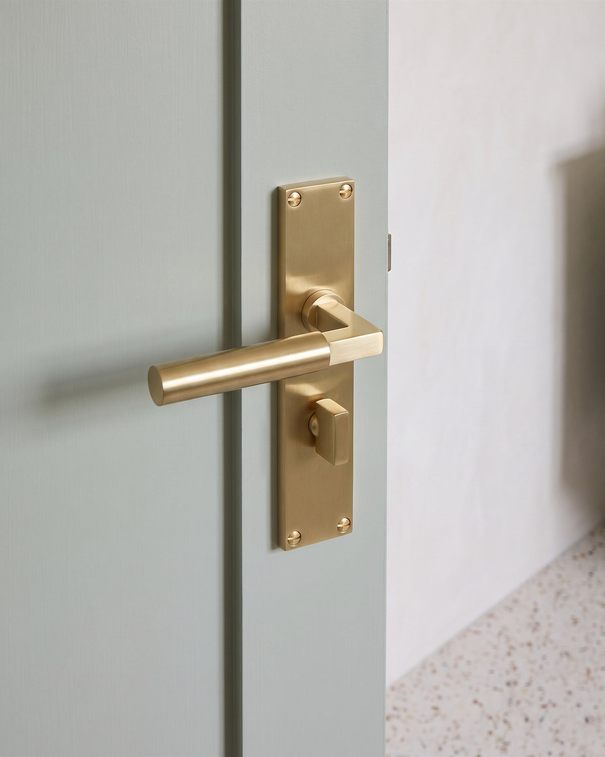 Satin brass Bauhaus bathroom handle with a straight bar lever and thumbturn on a soft green-grey bathroom door