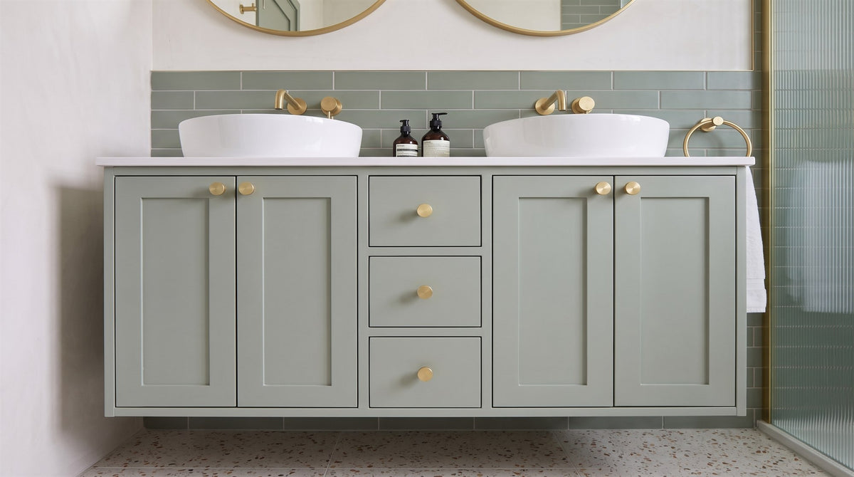 Green-grey double vanity with round satin brass mirrors and knurled satin brass cabinet knobs above a terrazzo floor