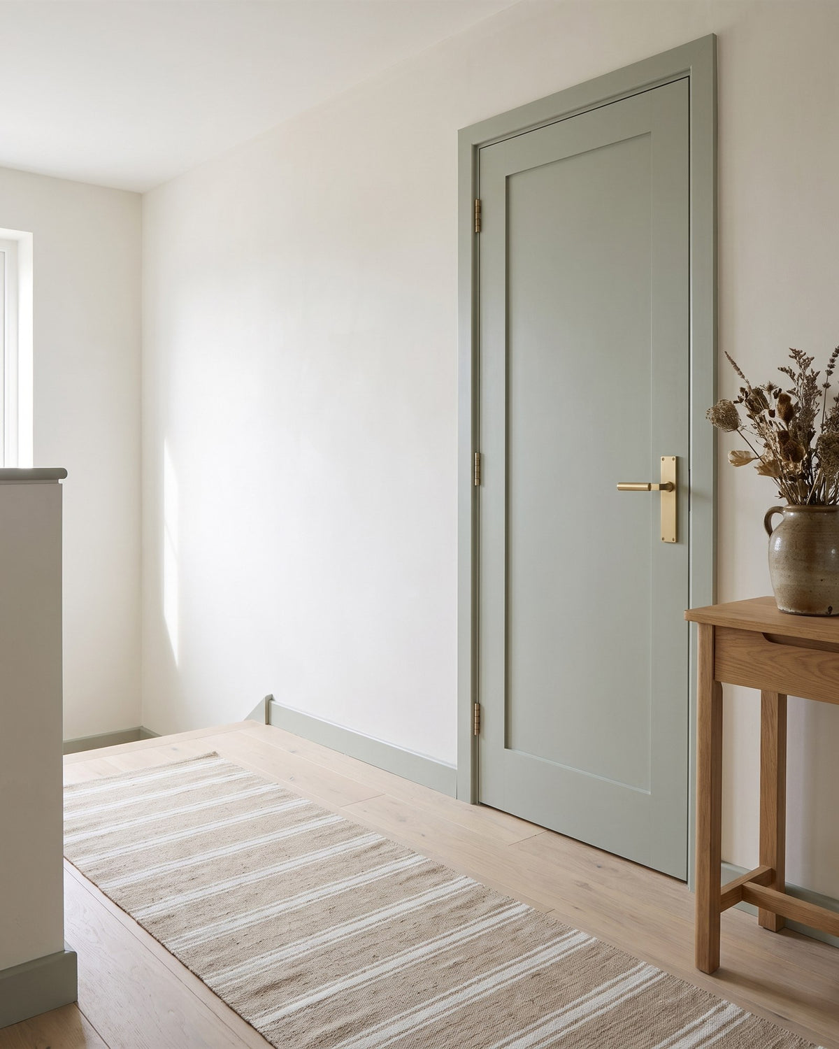 Green-grey bedroom door on a bright landing with a satin brass Bauhaus latch handle, an oak console and a striped runner