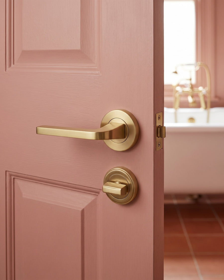 Satin brass Bellagio lever and stepped turn and release on a clay pink bathroom door, roll-top bath beyond