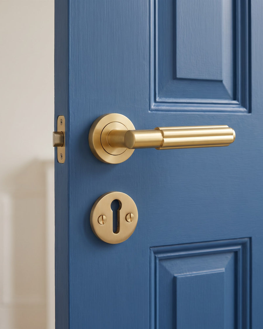Satin brass Bloom lever on rose with a satin brass key escutcheon directly below it, near the opening edge of a mid blue panelled door