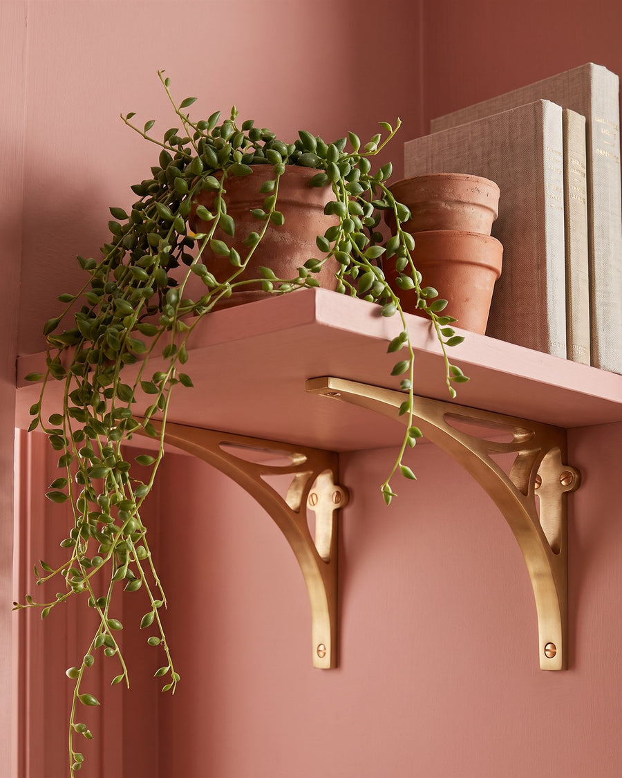 Satin brass bridge shelf brackets holding a clay pink shelf with terracotta pots and a trailing plant