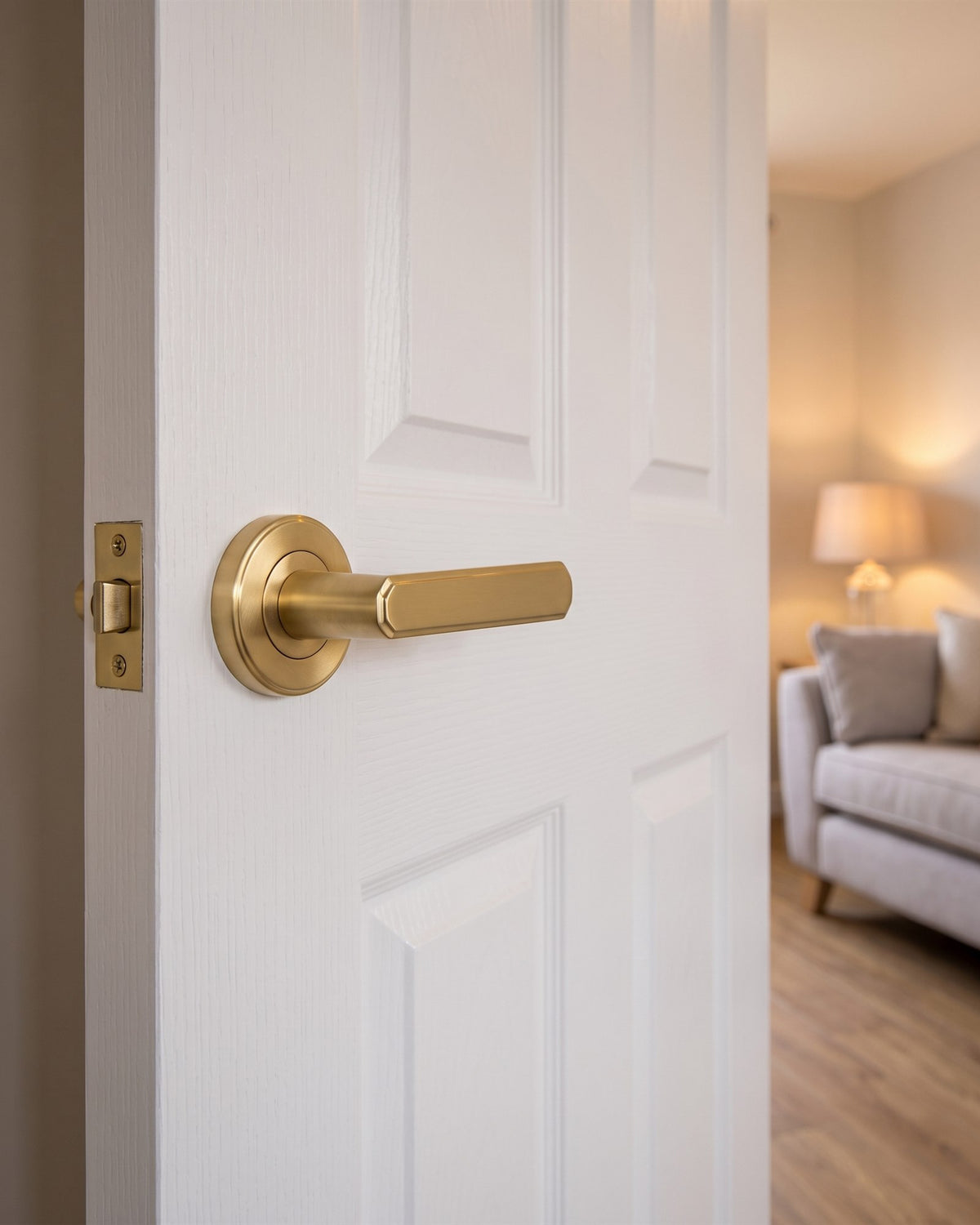 Satin brass Century lever on rose on a white moulded living room door, with a sofa and lamp beyond