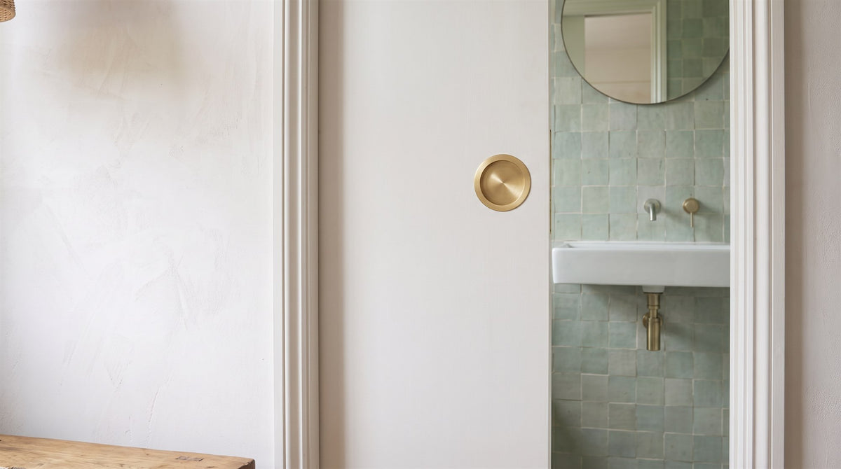 Satin brass circular flush pull set into a white pocket door sliding open onto a green tiled en suite