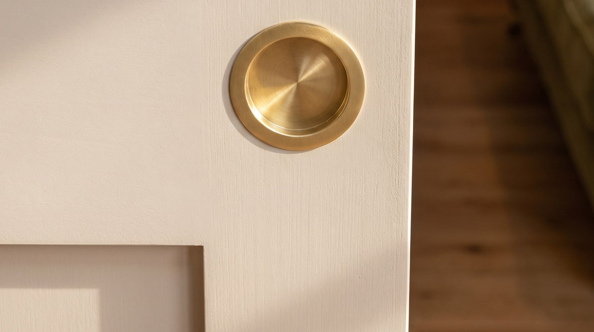 Circular satin brass flush pull set into a warm putty painted pocket door in soft afternoon light