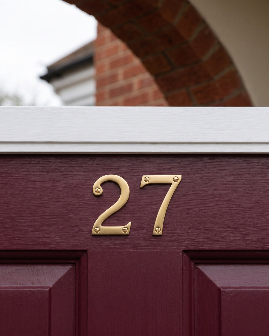 Satin brass door numbers 27 screwed to the top rail of a burgundy front door under a brick arch