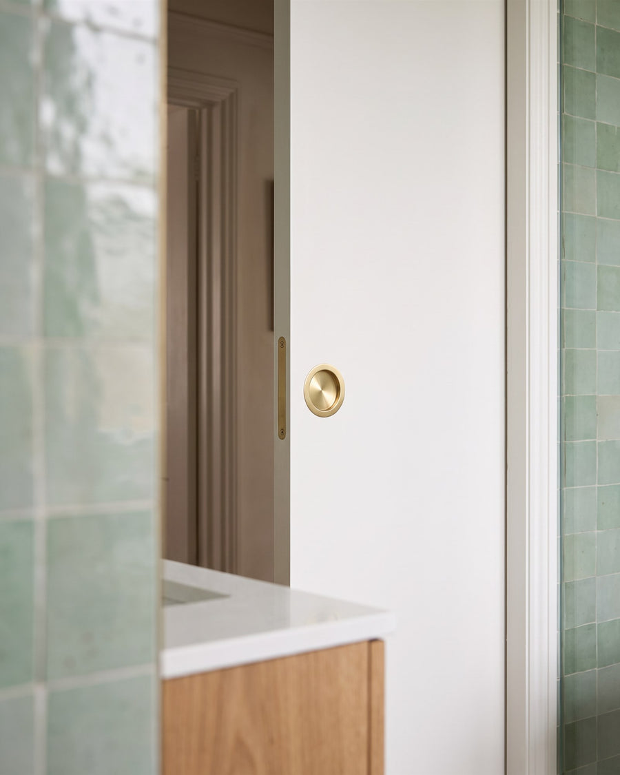 Satin brass circular flush pull on the en suite side of a white pocket door, green zellige tiles in the foreground