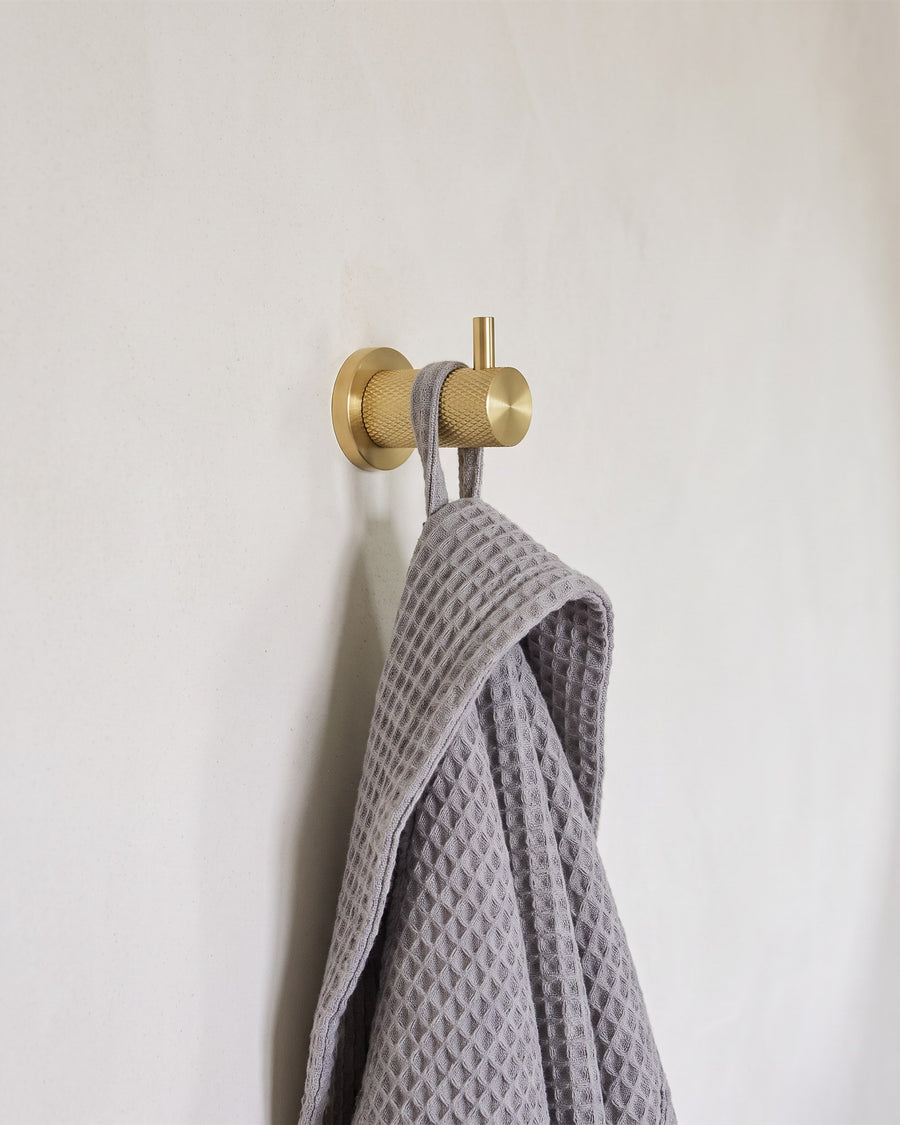 Satin brass diamond-knurled coat hook holding a grey waffle robe on a warm white plaster wall