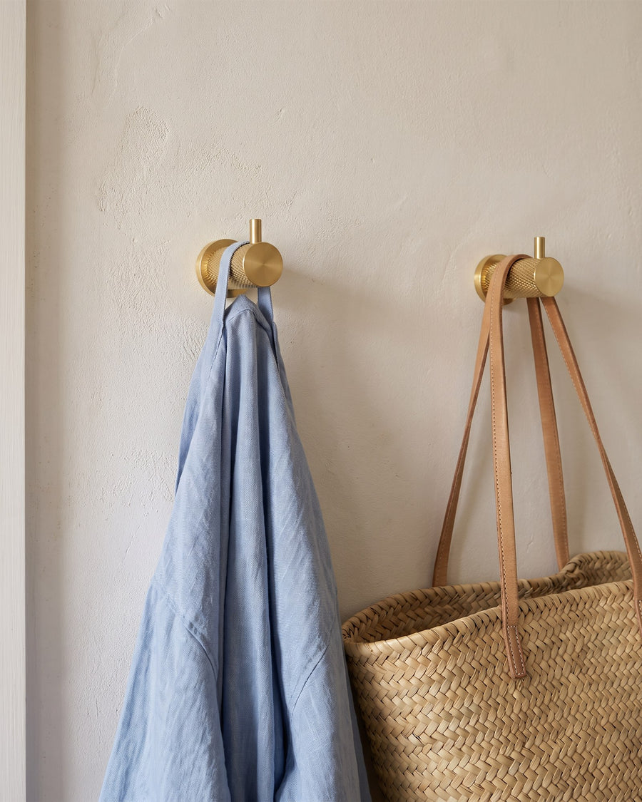 Two satin brass knurled coat hooks holding a blue linen robe and a woven straw bag