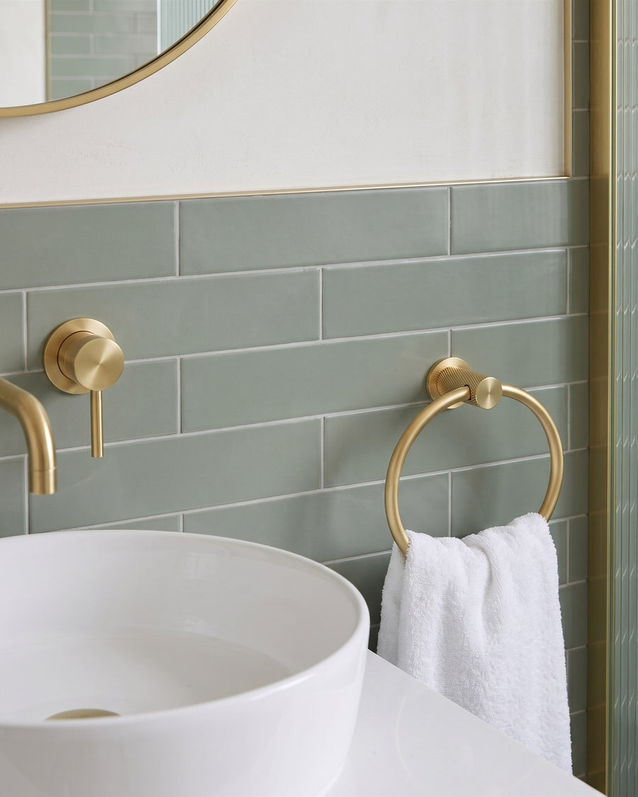 Satin brass knurled towel ring holding a white hand towel on green-grey metro tiles beside a white basin