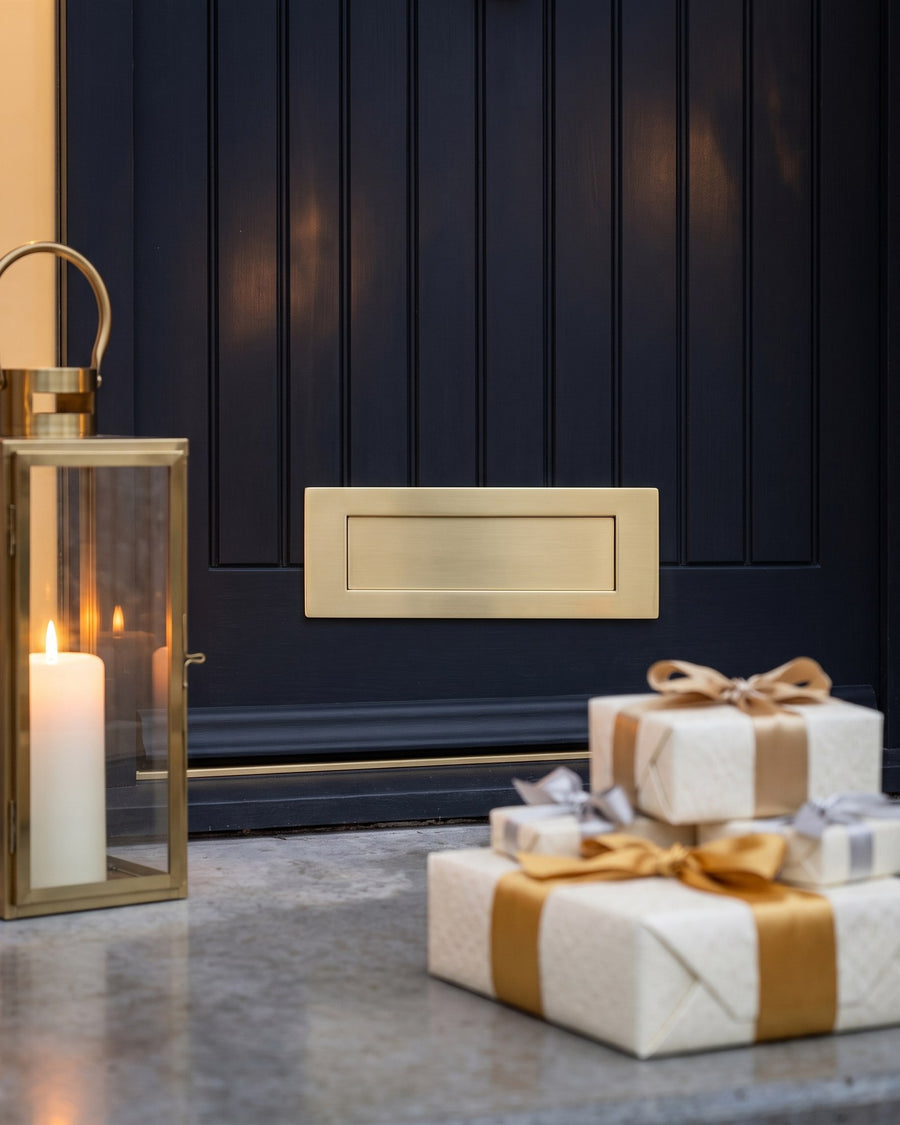 Satin brass letter plate on a matt black boarded door with ivory gifts tied in gold and silver ribbon and a brass lantern on the concrete step