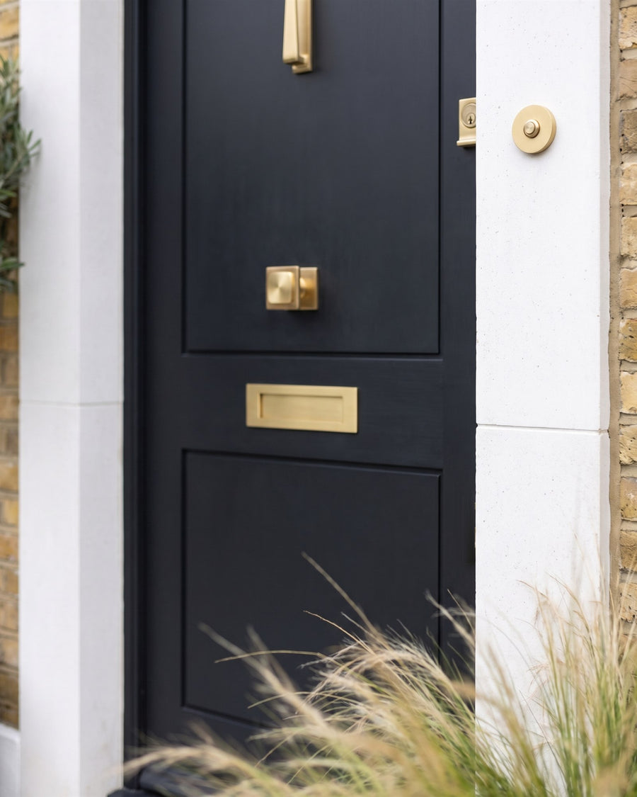 Satin brass square centre knob, letter plate and small cylinder pull on a near-black door, with a round satin brass bell push on the white stone surround