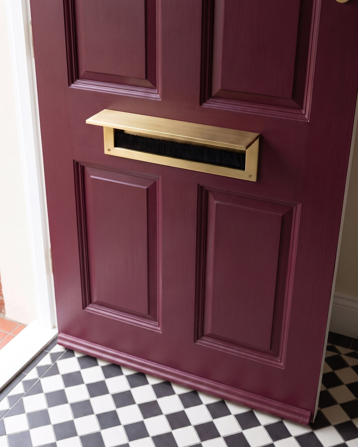 Inside of a burgundy front door with a satin brass letter tidy and brush draught excluder above a chequerboard floor