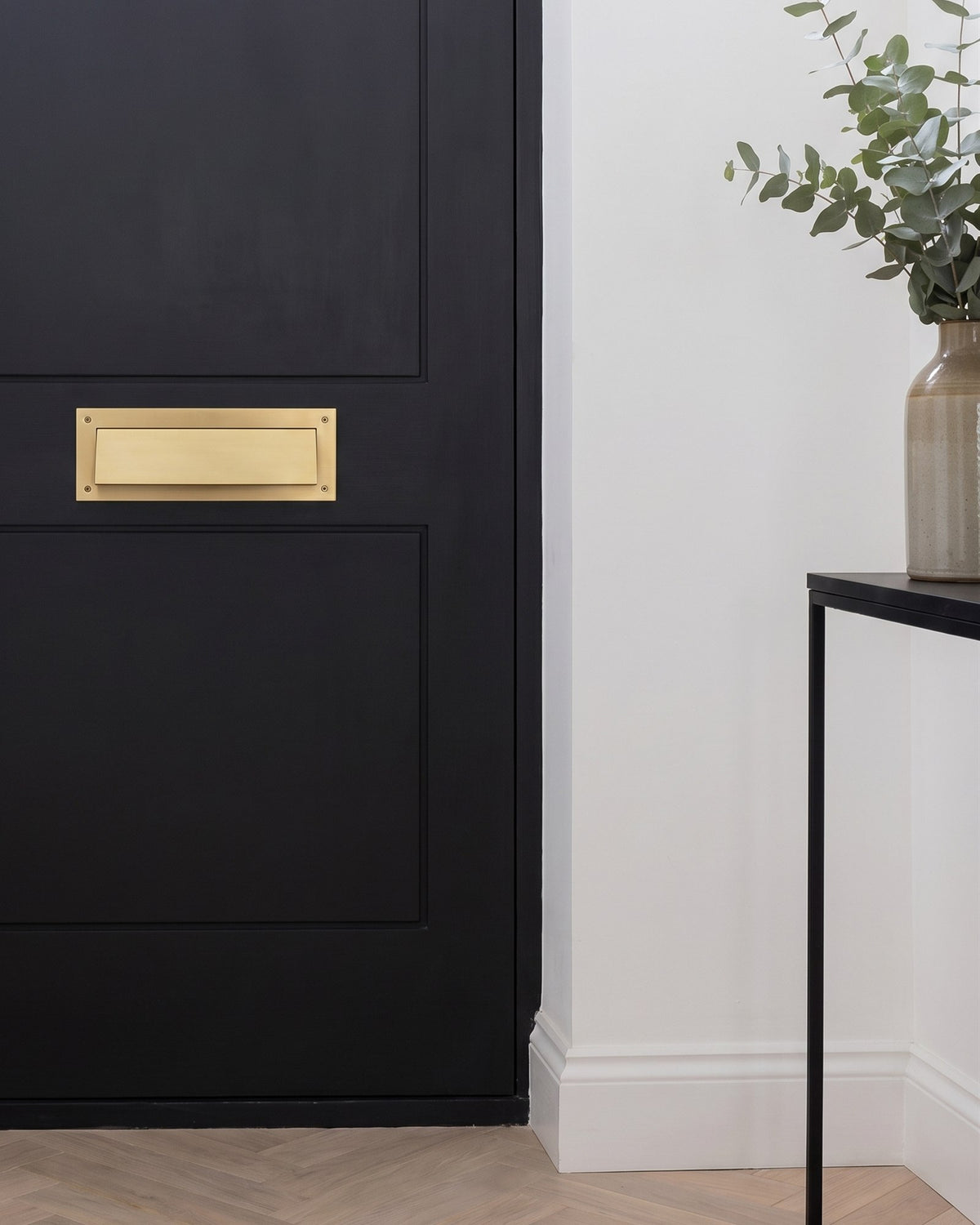 Satin brass letter tidy with draught excluder on the inside of a near-black front door, with a herringbone oak floor and a vase of eucalyptus
