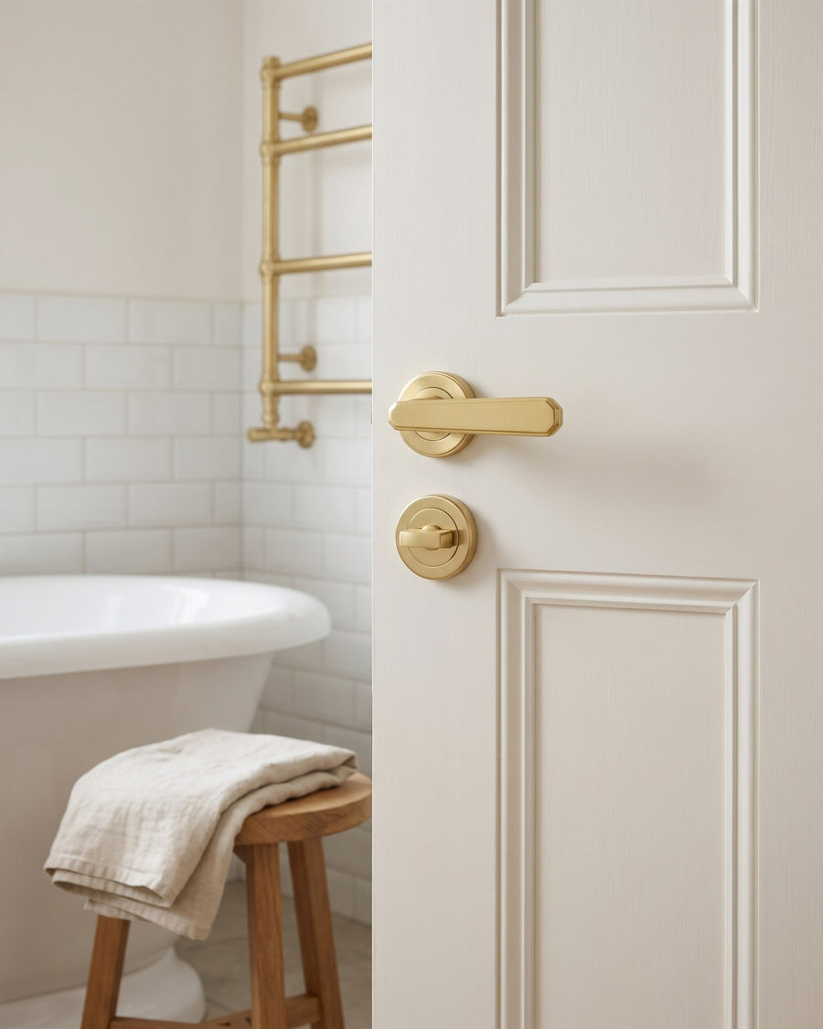 Bathroom door with a satin brass lever and matching turn and release, a roll-top bath and brass towel rail beyond