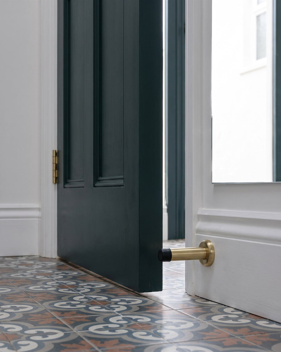 Inky green door resting on a satin brass projection door stop on white skirting, a satin brass hinge and patterned floor tiles