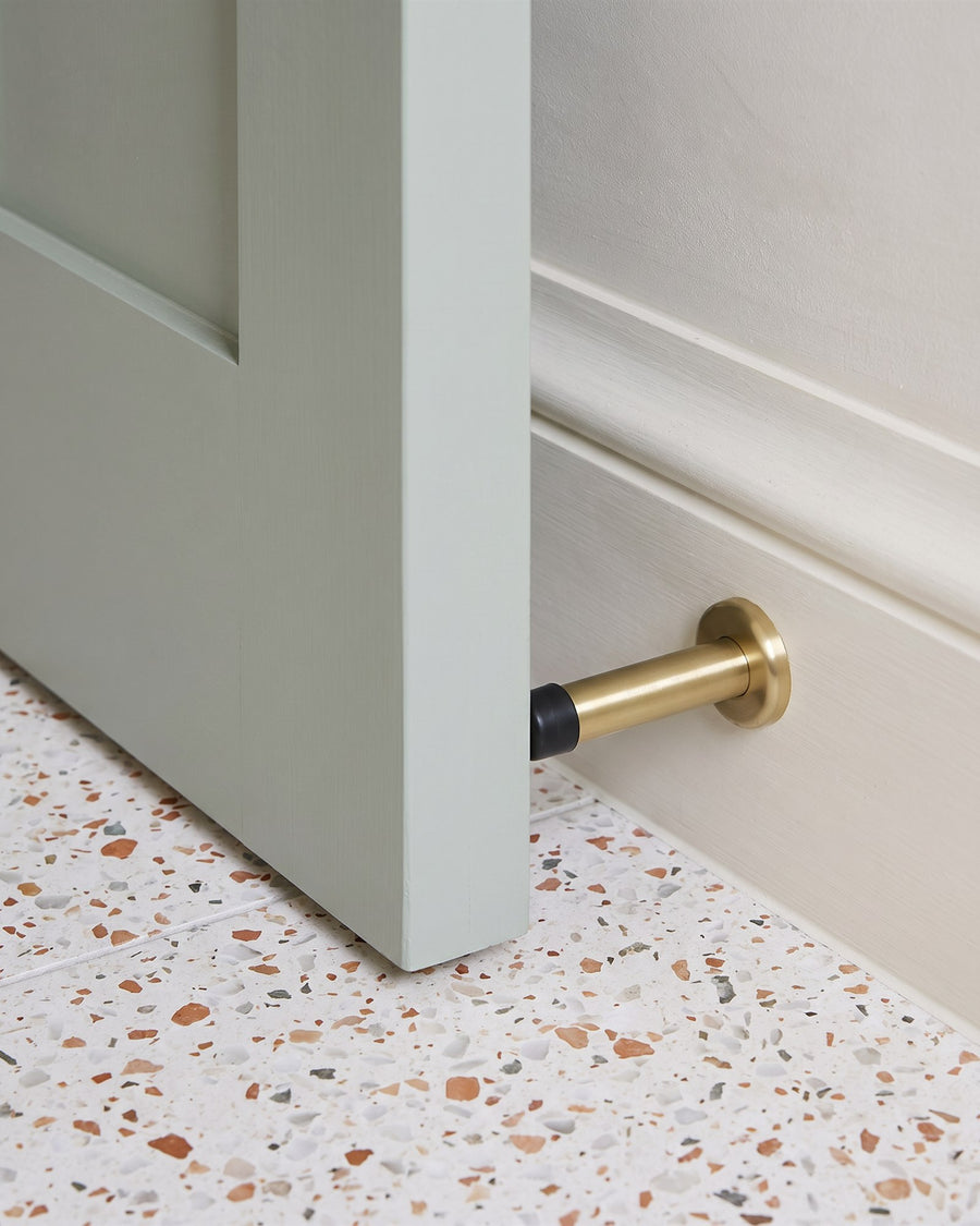 Green-grey bathroom door resting against a satin brass projection door stop on the skirting above a terrazzo floor