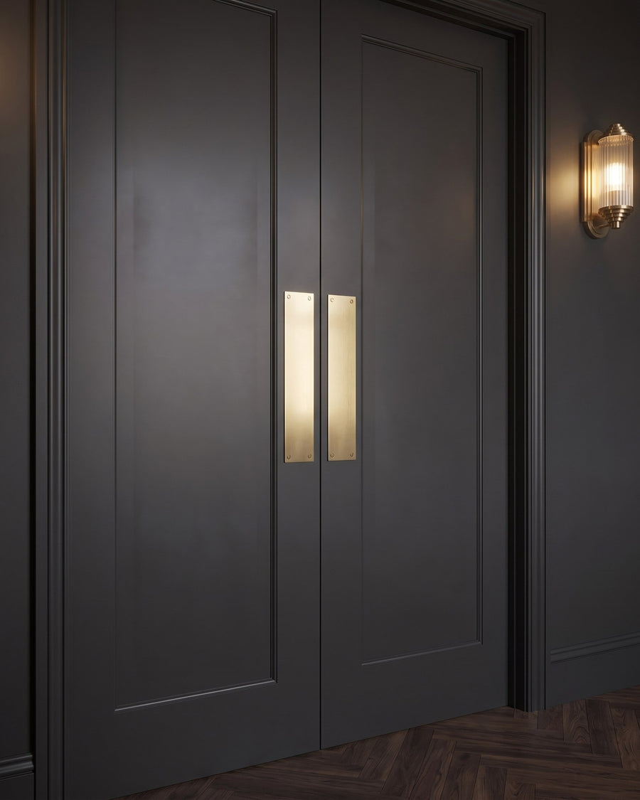 Pair of charcoal push-open double doors with a plain satin brass push plate on each leaf, lit by a ribbed-glass brass wall light