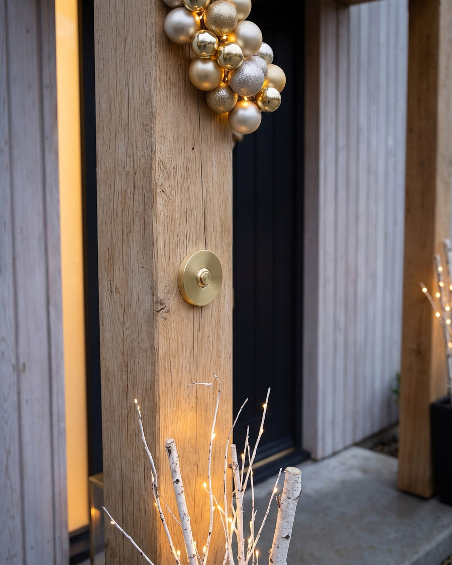 Round satin brass bell push on a chunky oak porch post beneath a cluster of gold and champagne baubles, with lit white birch branches below