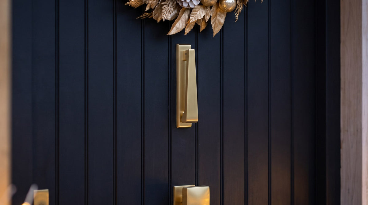 Satin brass slim door knocker above a square satin brass centre knob on a matt black vertical-boarded front door, with the edge of a gilded leaf wreath above