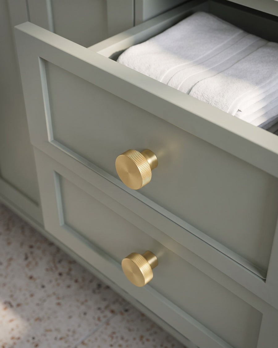Satin brass Thaxted line-knurled cabinet knobs on green-grey shaker vanity drawers, one drawer open with folded towels