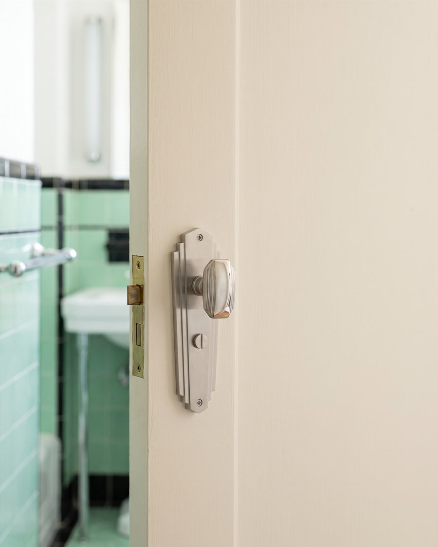 Satin chrome Charlston Art Deco bathroom knob set with turn on a cream door, mint and black tiled bathroom beyond
