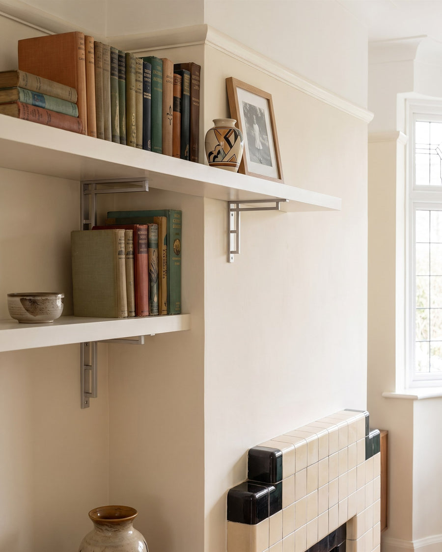 Satin chrome Art Deco shelf brackets under white alcove shelves holding old books beside a 1930s tiled fireplace