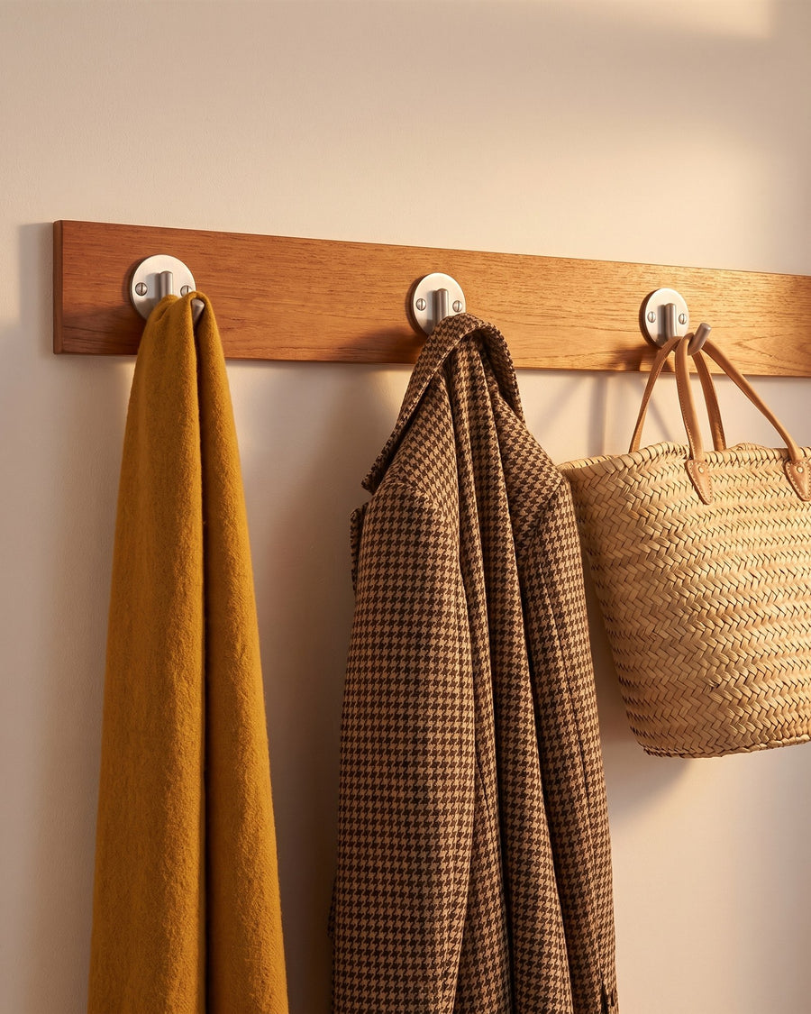 Satin chrome coat hooks on round plates along a teak batten holding a mustard scarf, houndstooth coat and basket