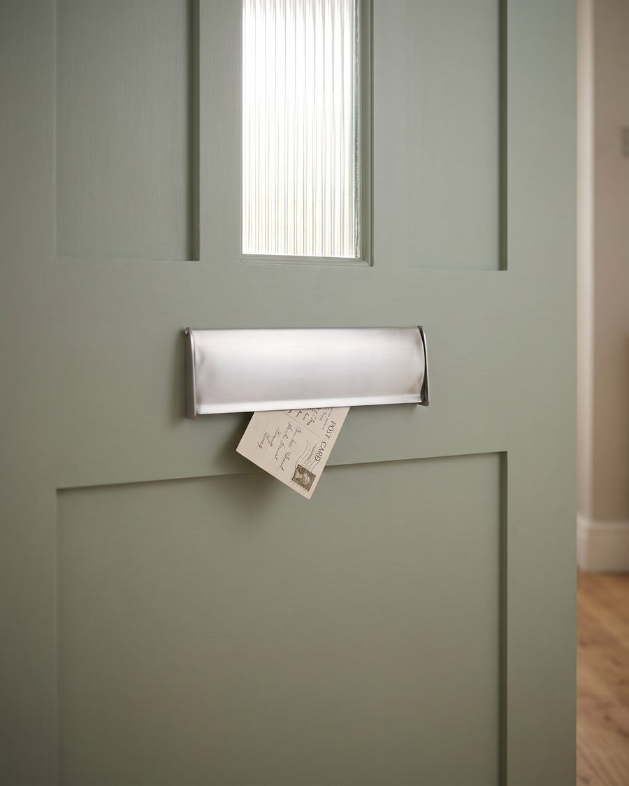 Satin chrome letter box tidy on the inside of a sage front door below the reeded glass panel, with a postcard just delivered