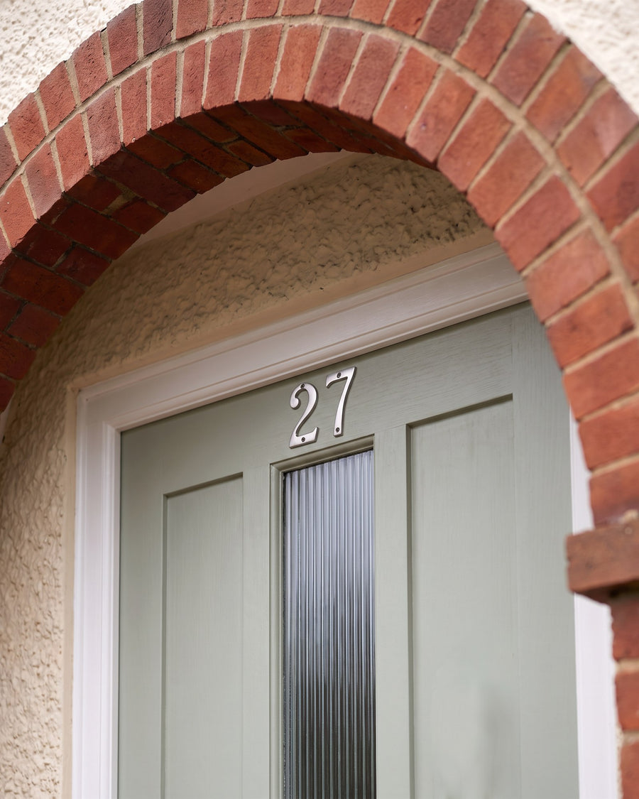 Satin chrome numbers 27 on a sage door beneath a red brick porch arch, above a reeded glass panel
