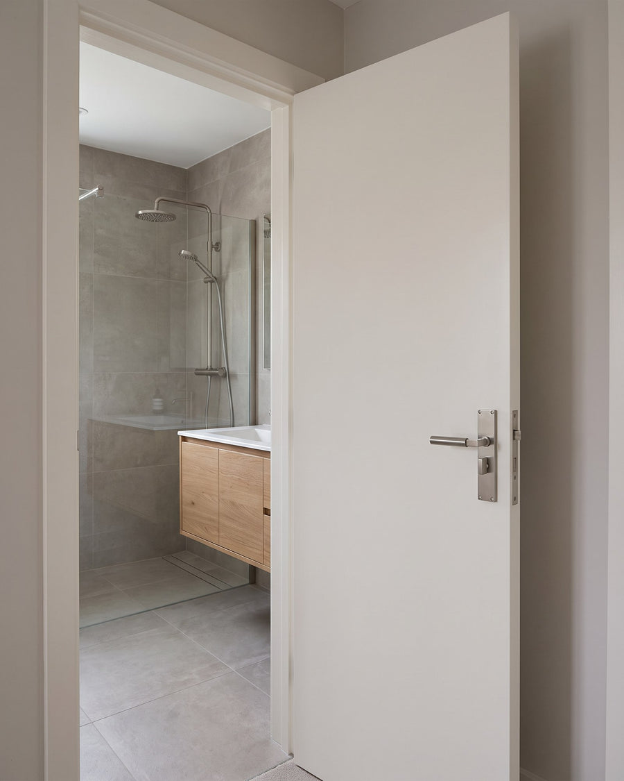 Warm white bathroom door with a satin nickel Bauhaus bathroom handle, a porcelain-tiled shower room and oak vanity beyond