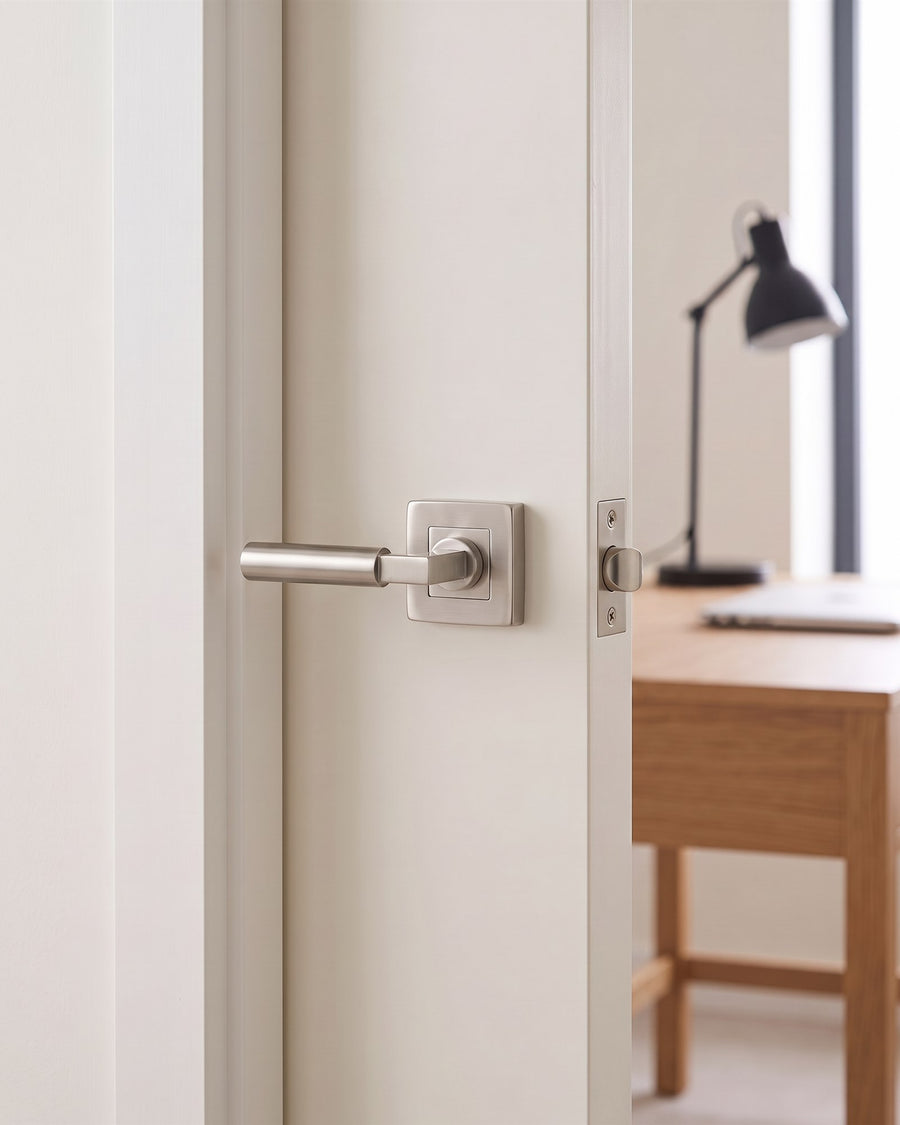 Satin nickel Bauhaus lever on a square rose on a warm white door, with a satin nickel latch in the door edge and an oak desk and black lamp beyond