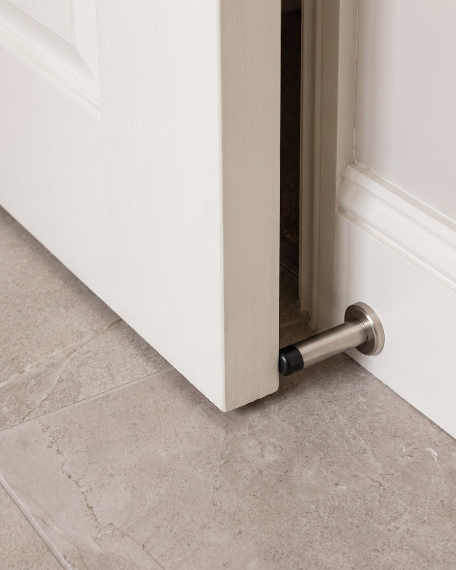 White bathroom door resting against a satin nickel cylinder door stop on the skirting above stone-effect floor tiles