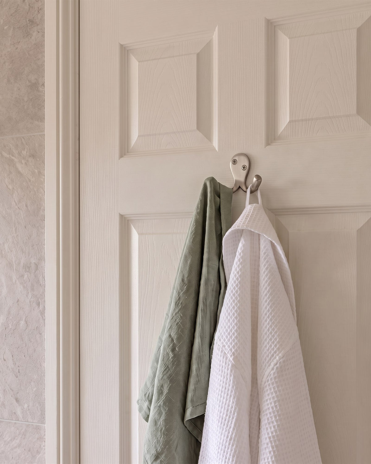 Satin nickel double coat hook holding a white waffle robe and a sage green hand towel on a white panelled bathroom door
