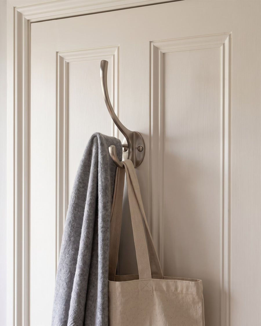 Satin nickel hat and coat hook holding a grey scarf and canvas bag on a warm white panelled door