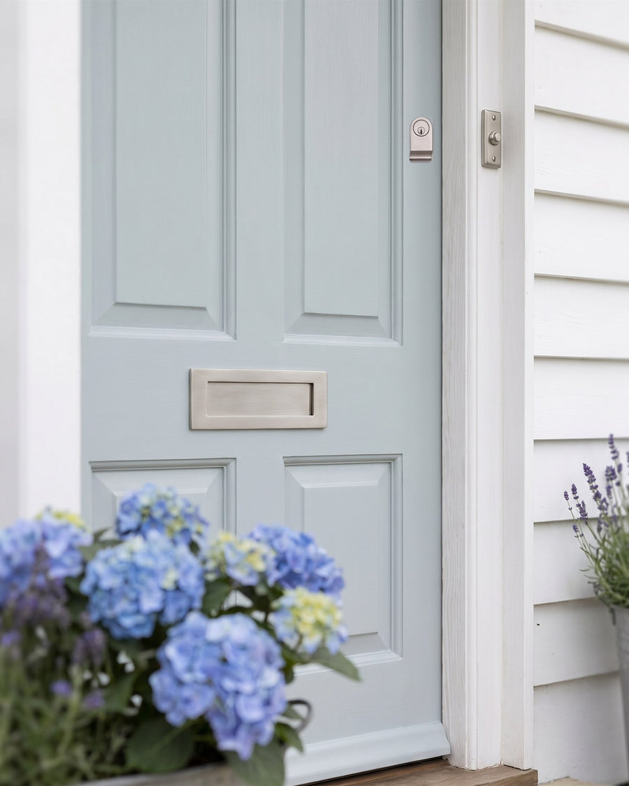 Satin nickel letter plate and cylinder pull on a powder blue front door, with a rectangular satin nickel bell push on the white frame and hydrangeas in front