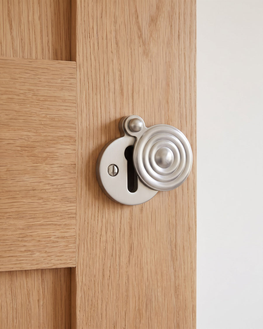 Satin nickel reeded covered escutcheon with its swivel cover turned aside on a natural oak veneer door