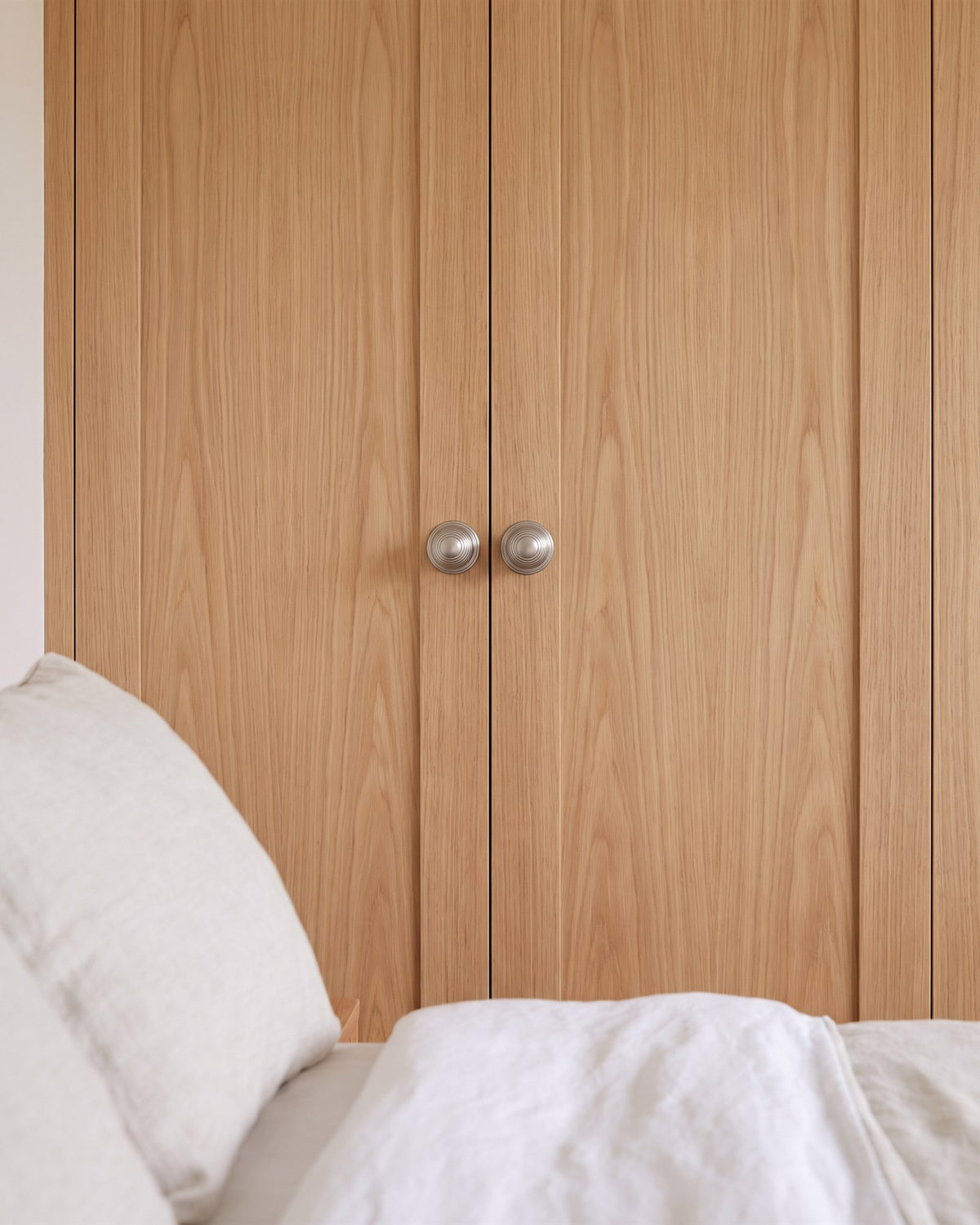 Pair of satin nickel reeded cupboard knobs on natural oak veneer fitted wardrobe doors beside a bed with white linen