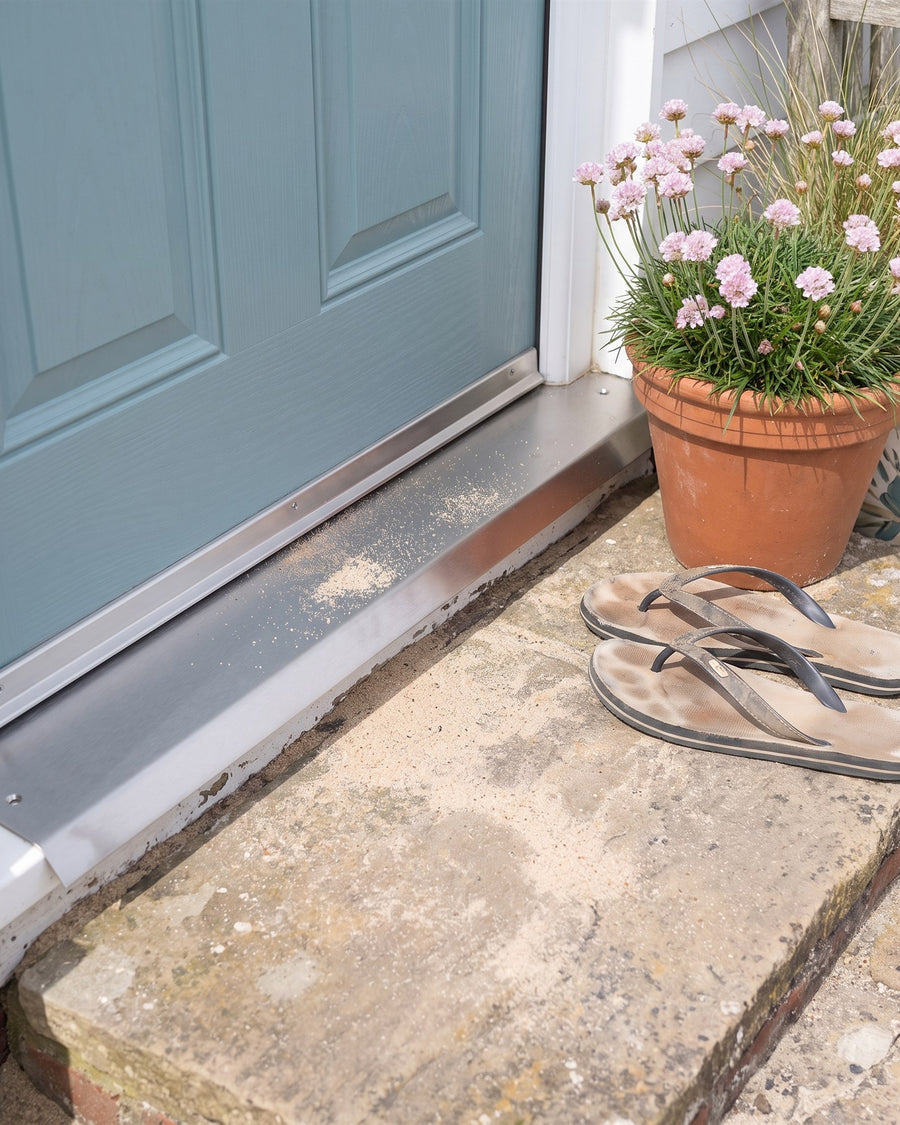 Satin stainless steel threshold strip on a sandy cottage doorstep with flip-flops and sea thrift