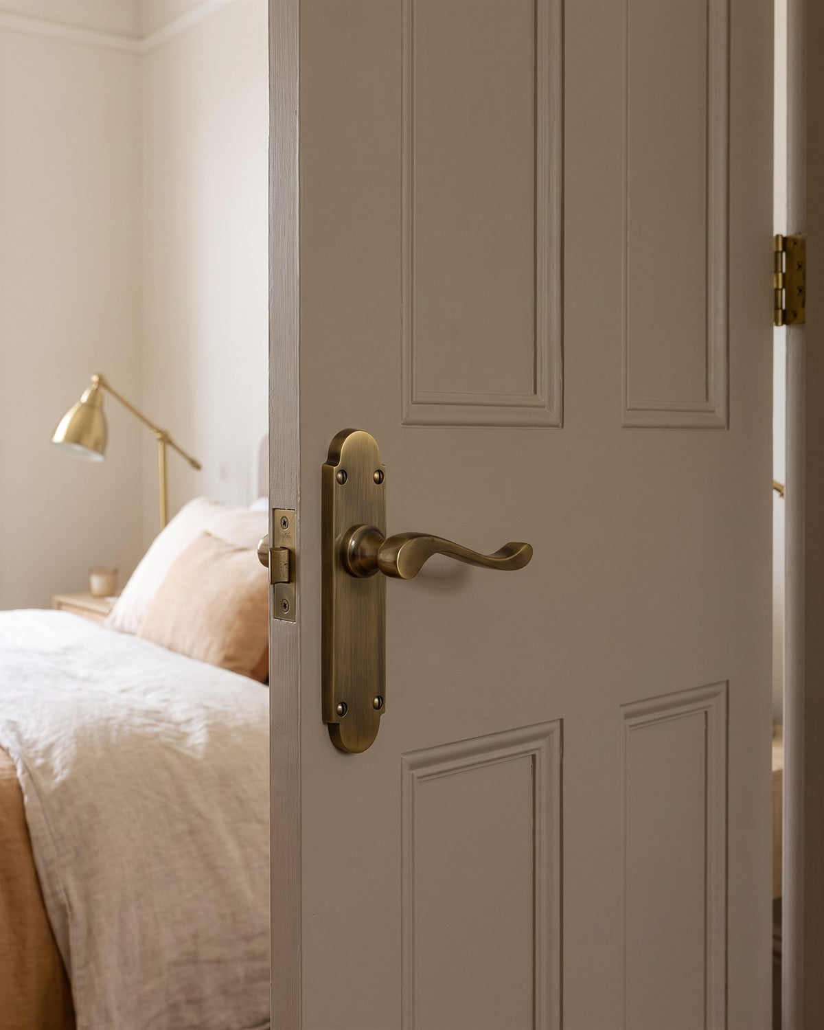Antique brass Savoy scroll lever on a warm putty bedroom door, linen bed and brass lamp beyond