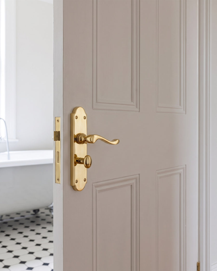 Polished brass Savoy scroll lever bathroom handle with turn on a white door, black and white tiled bathroom beyond