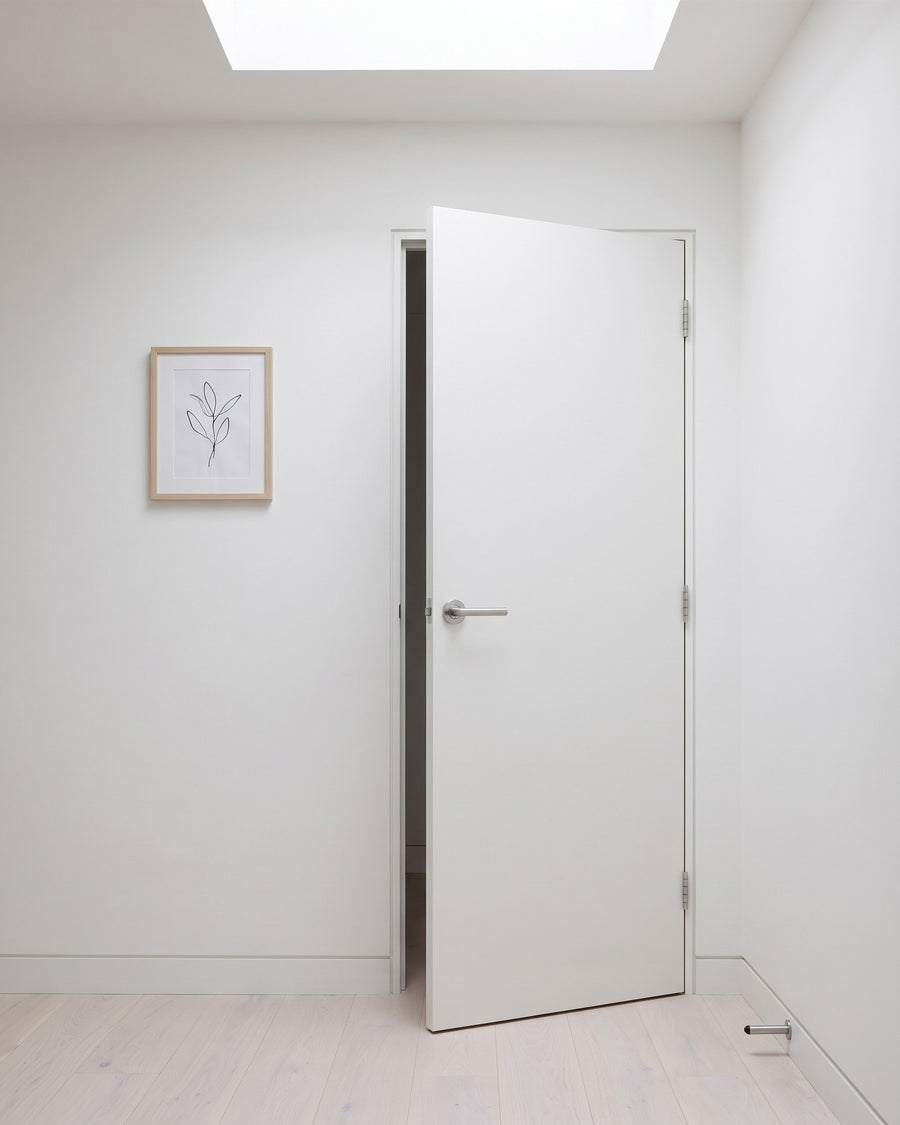 Full-height flush white bedroom door ajar on a calm landing with a roof light, fitted with a satin chrome T-bar lever on rose