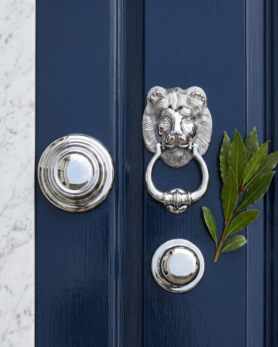 Polished chrome Sloane centre knob, lion head knocker and round centre knob laid on a navy panel beside marble and bay leaves