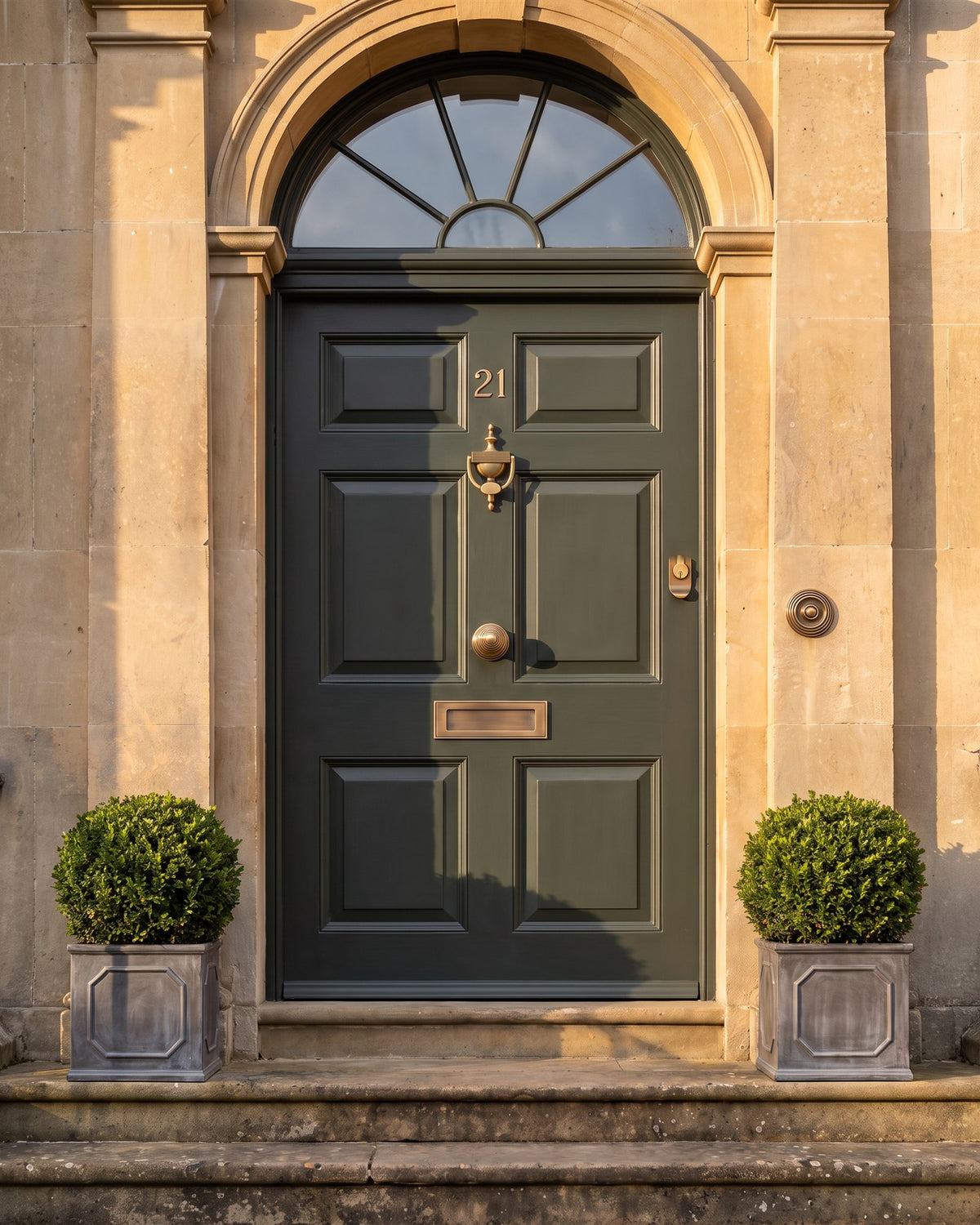 Smoky green front door in a Bath stone doorcase in late-afternoon sun, the reeded antique brass knocker, knob, letter plate and bell push catching the light