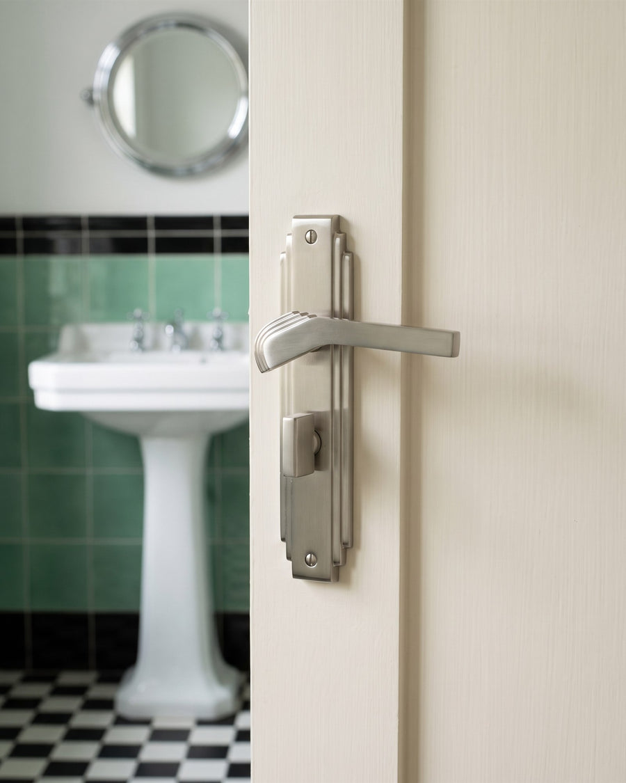 Satin nickel Tiffany bathroom handle with thumbturn on a cream door, jade and black tiles and a pedestal basin beyond