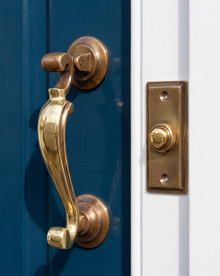 Unlacquered brass profile doctor knocker on a navy door and stepped bell push on a white doorcase, polished bright where hands touch and mellowed elsewhere