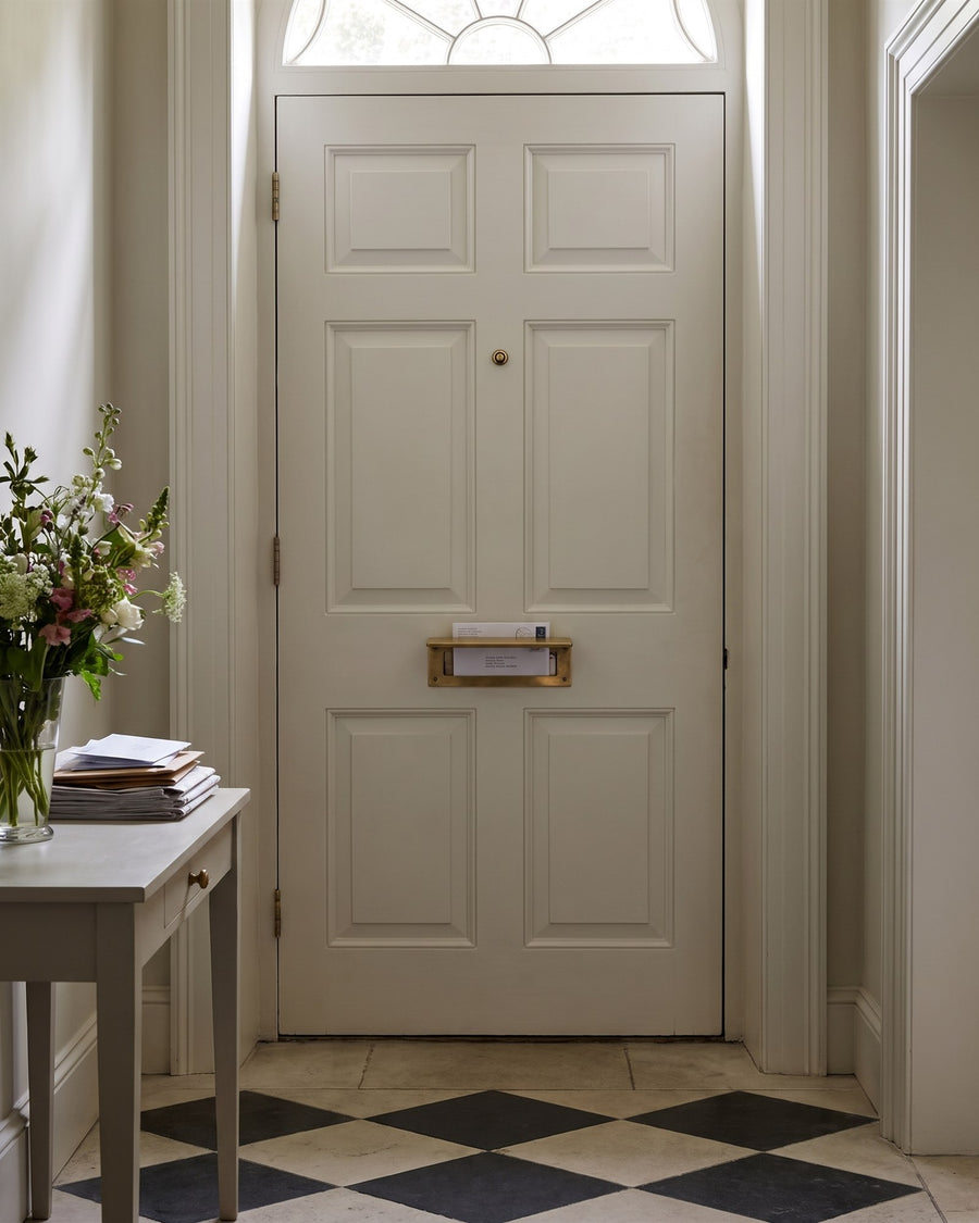Inside of an off-white Georgian front door with an unlacquered brass letter tidy above a black and white chequered stone hall floor