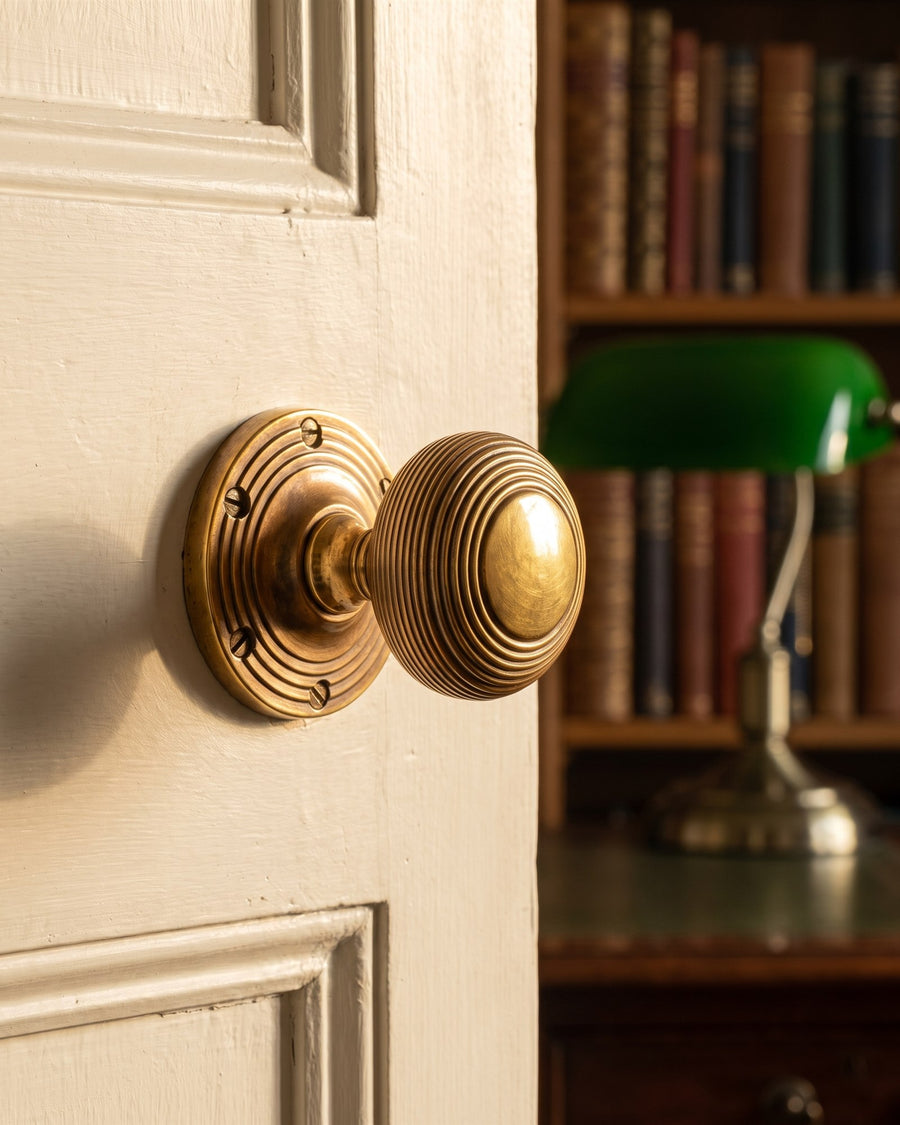 Unlacquered brass reeded mortice knob on a painted panelled study door, with darker grooves and bright worn edges, bookshelves beyond