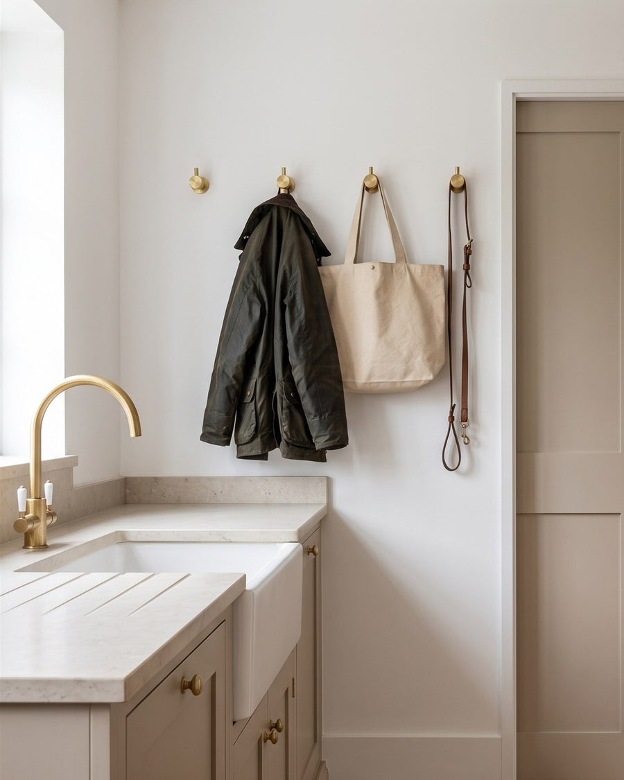 Utility room with a butler sink, satin brass tap and a row of satin brass coat hooks holding a waxed jacket, tote bag and dog lead beside a putty pocket door